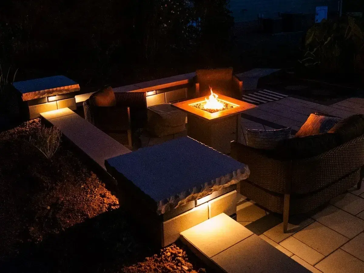 Evening view of a stone seating area with lit seat walls around a fire table